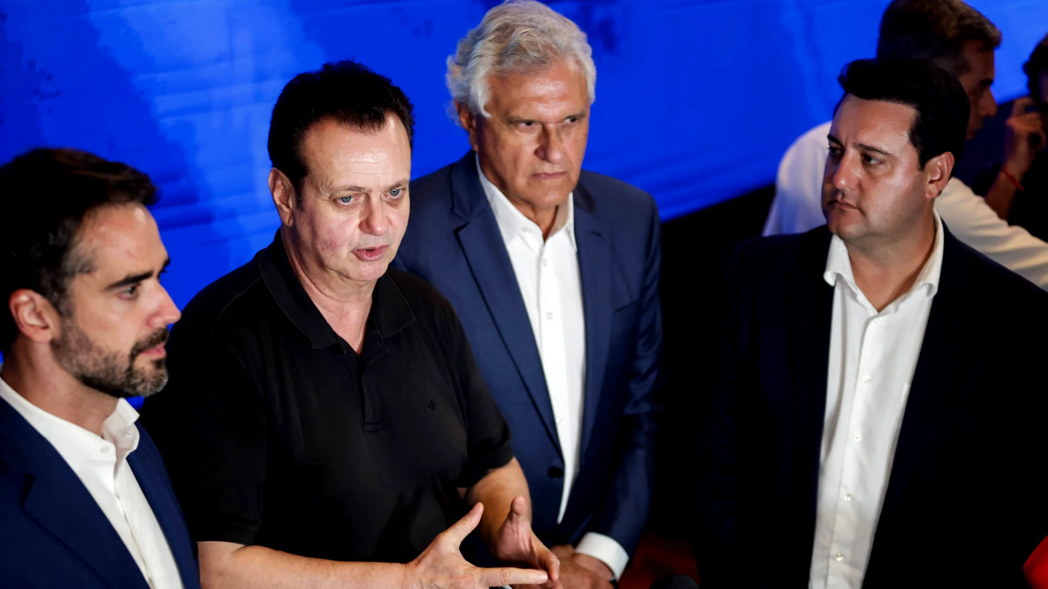 A imagem mostra quatro figuras políticas brasileiras em um ambiente interno. Da esquerda para a direita: Eduardo Leite, de perfil, vestindo paletó azul e camisa branca; Gilberto Kassab, ao centro, vestindo uma camisa polo preta e gesticulando com as mãos enquanto fala; Ronaldo Caiado, ao fundo, de terno azul e cabelos brancos; e Ratinho Júnior, à direita, vestindo paletó escuro e camisa branca, observando a fala. À frente deles, aparecem microfones de imprensa. O fundo é composto por um painel azul iluminado.