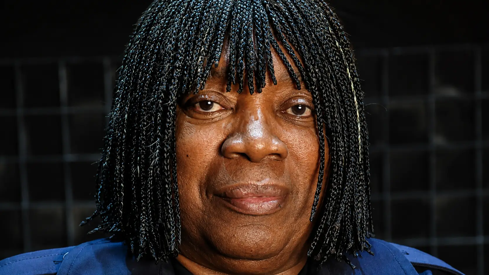 Retrato em close do cantor Milton Nascimento, com tranças e roupa azul, diante de um fundo escuro.