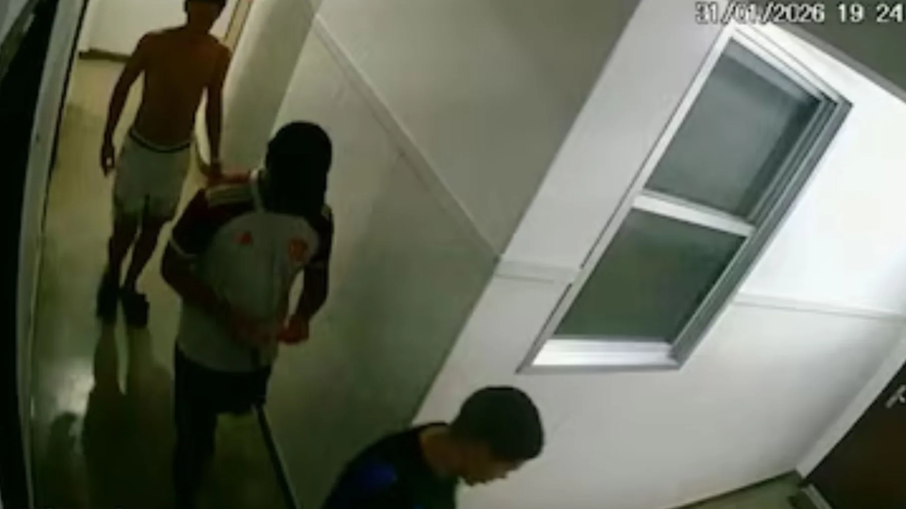 Imagem de câmera de segurança (CCTV) em um corredor de prédio. Três jovens são vistos caminhando pelo corredor em direção a um apartamento. No canto superior direito, o registro de data e hora marca 31/01/2026 às 19:24. Um dos jovens está sem camisa e os outros dois vestem camisetas escuras.