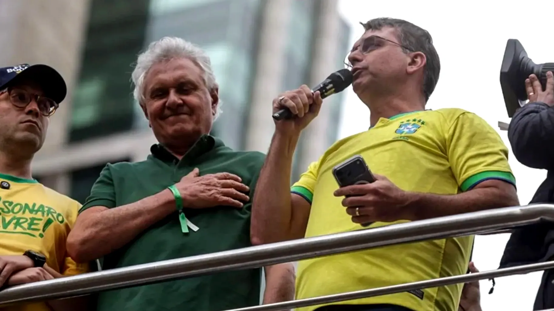 Flávio Bolsonaro discursa em microfone durante ato na Avenida Paulista, ao lado do governador Ronaldo Caiado e apoiador com camiseta escrito "Bolsonaro Livre!".