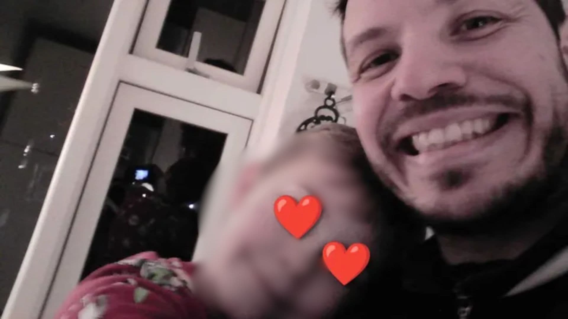 Alexander Rocha sorri ao lado de seu filho; a criança está encostada em seu ombro e tem o rosto protegido por dois emojis de coração vermelho.