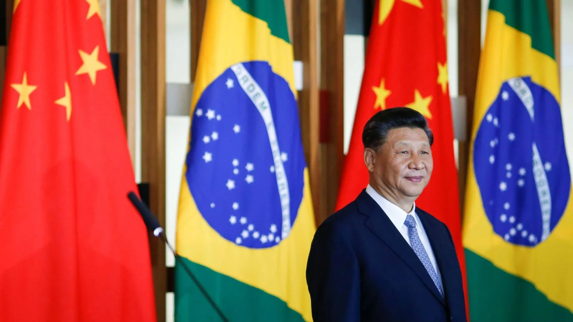 Xi Jinping sorrindo à frente das bandeiras da China e do Brasil.