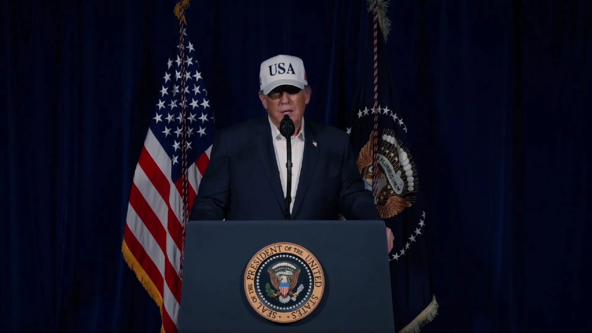 : O presidente Donald Trump posicionado atrás de um púlpito azul com o selo oficial da Presidência dos Estados Unidos. Ele veste um terno escuro, camisa branca e um boné branco com as iniciais "USA" em azul. Ao fundo, uma cortina azul escura ladeada pela bandeira dos Estados Unidos à esquerda e a bandeira presidencial à direita.